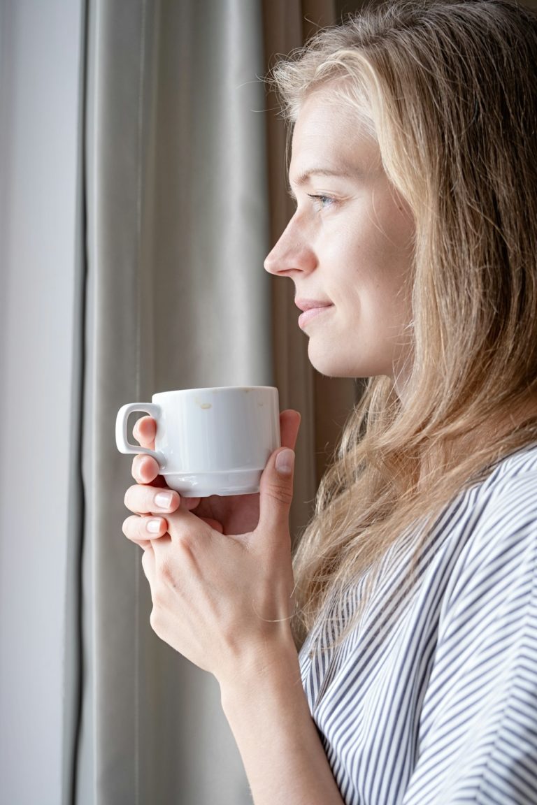 Happy woman standing by the window at hotel room drinking coffee